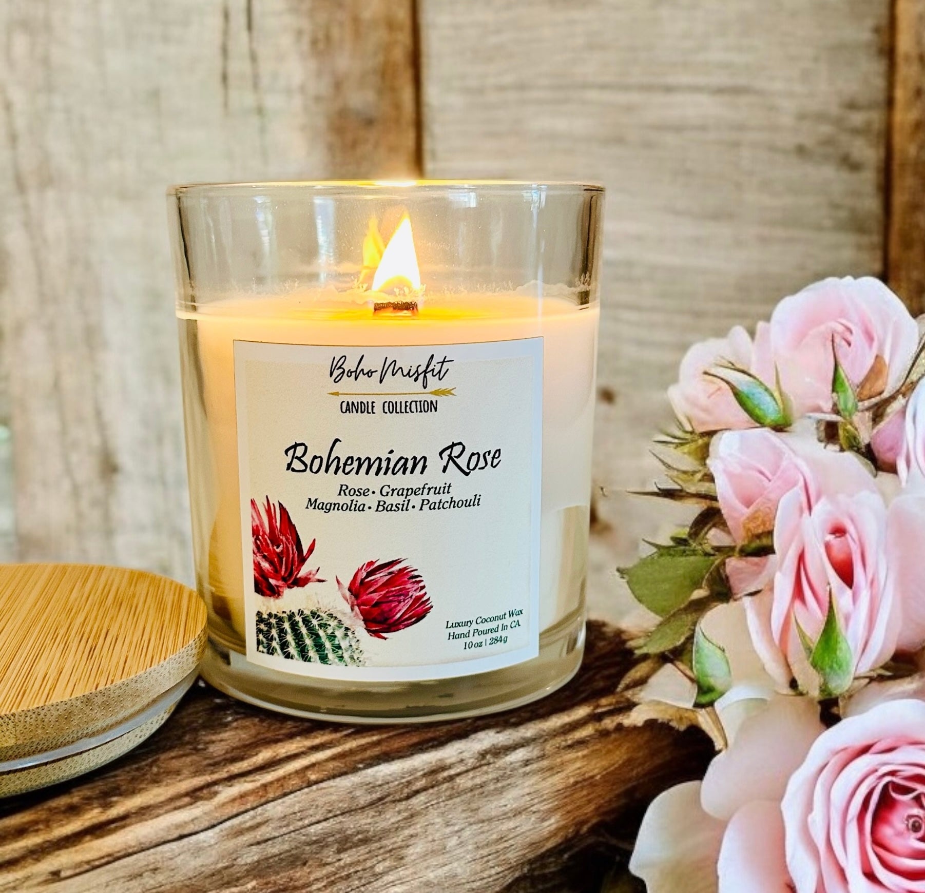 A coconut wax candle labeled 'Bohemian Rose' with a wooden wick, decorated with floral illustrations and burning on a wooden surface, alongside pink roses.